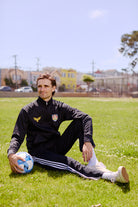 A man wearing the WC x SFP Zip-Up Jacket sits on grass with a soccer ball under the sun, capturing the US Men's National Team spirit in a park.