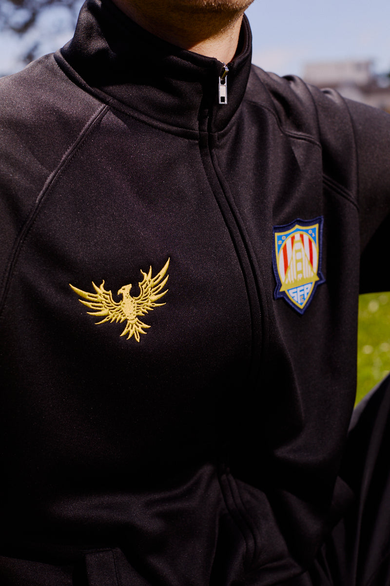 Wearing the WC x SFP Zip-Up Jacket—a black zip-up with a gold eagle emblem and a striking San Francisco Rising Phoenix shield patch on the chest.