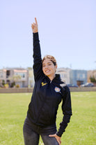 Woman standing on grass, smiling and raising one arm with index finger pointing to the sky, wearing the WC x SFP Zip-Up Jacket.
