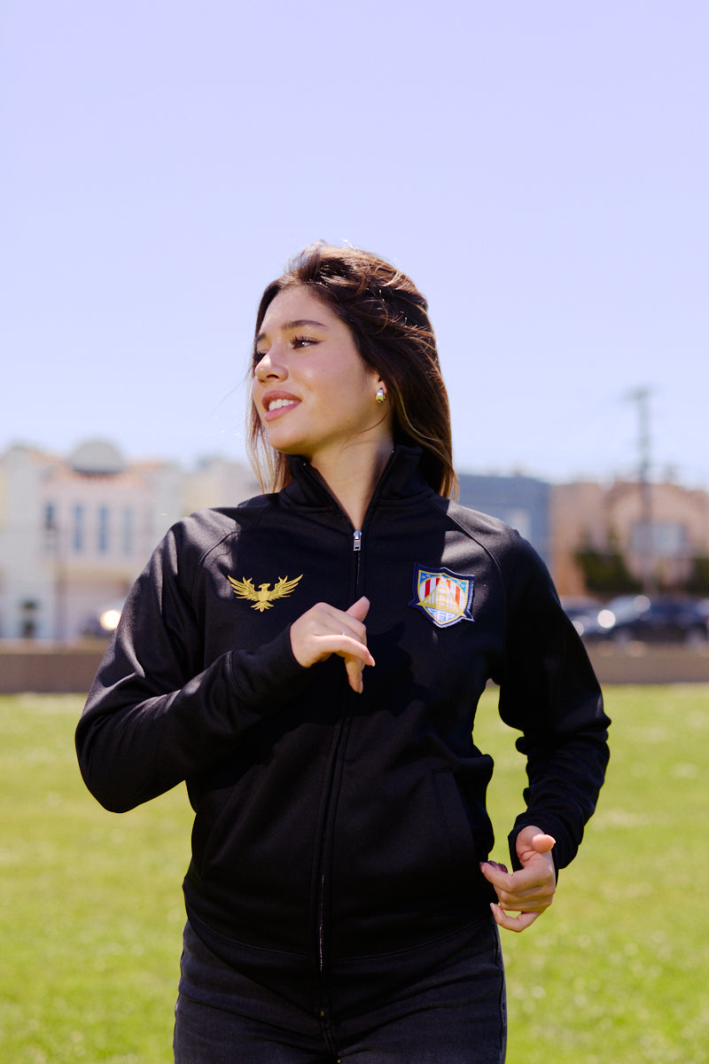 A woman runs outdoors on a sunny day wearing the WC x SFP Zip-Up Jacket, with city buildings and green grass in the background.