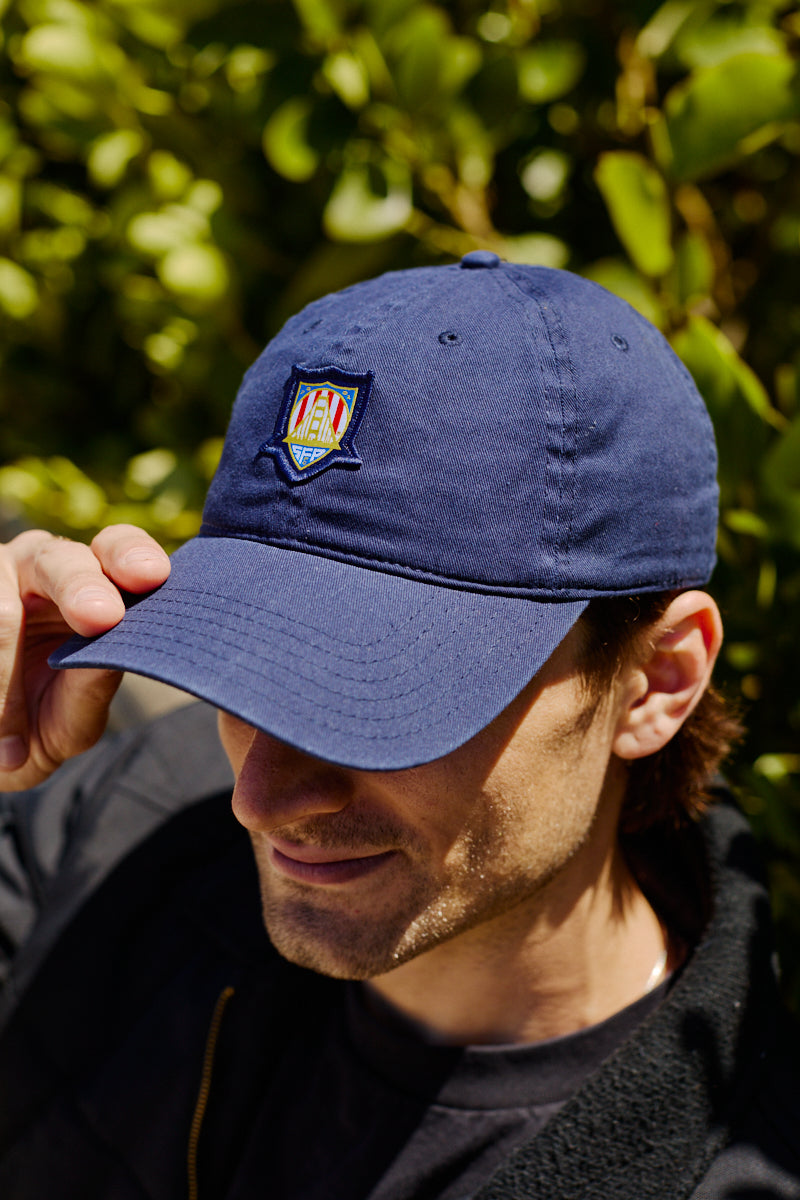 A man outdoors adjusts the brim of a blue WC x SFP Patched Hat, which features a colorful embroidered patch.