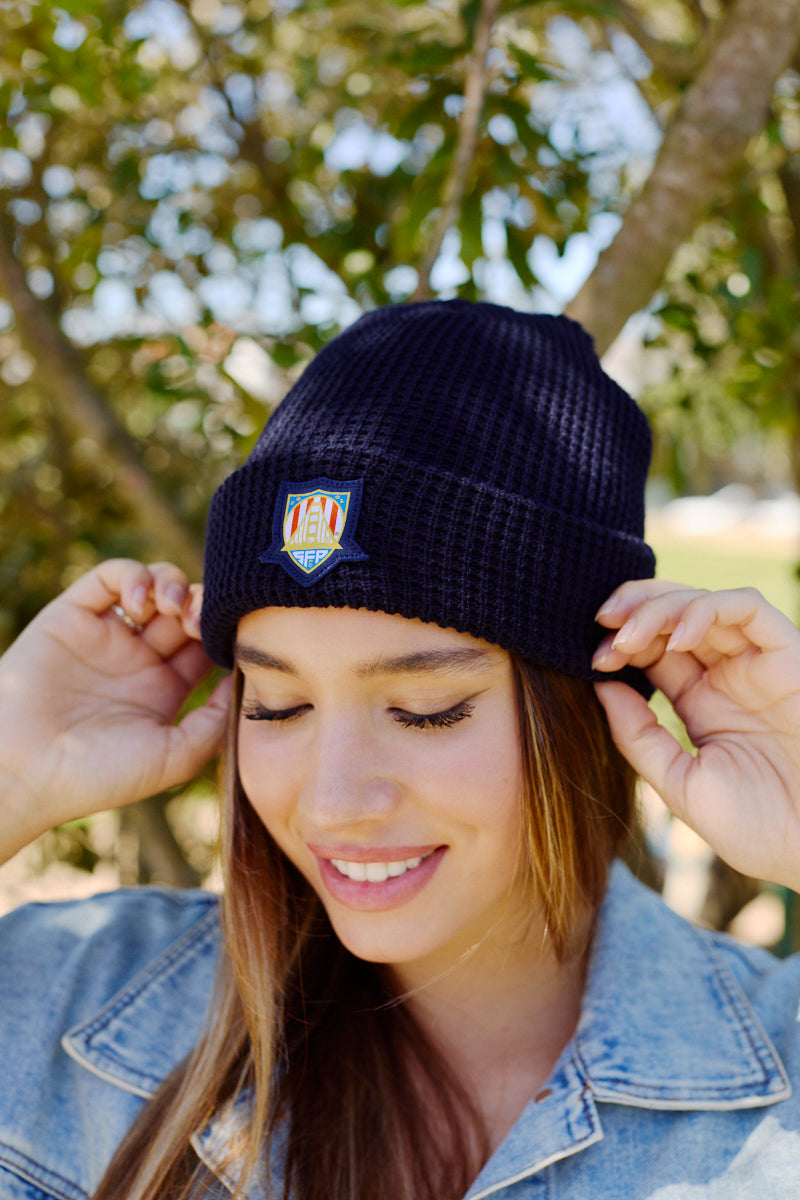 A woman smiles outdoors, wearing the WC x SFP Beanie with a colorful patch and a light denim jacket—the perfect look for cheering on the US Men's National Team.