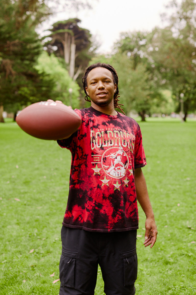 A man stands on grass in a park, holding a football out in front of him, wearing the Gold Rush Tee - Red Crystal Dye.