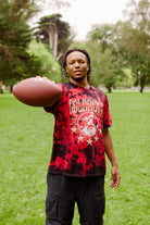 A man stands on grass in a park, holding a football out in front of him, wearing the Gold Rush Tee - Red Crystal Dye.