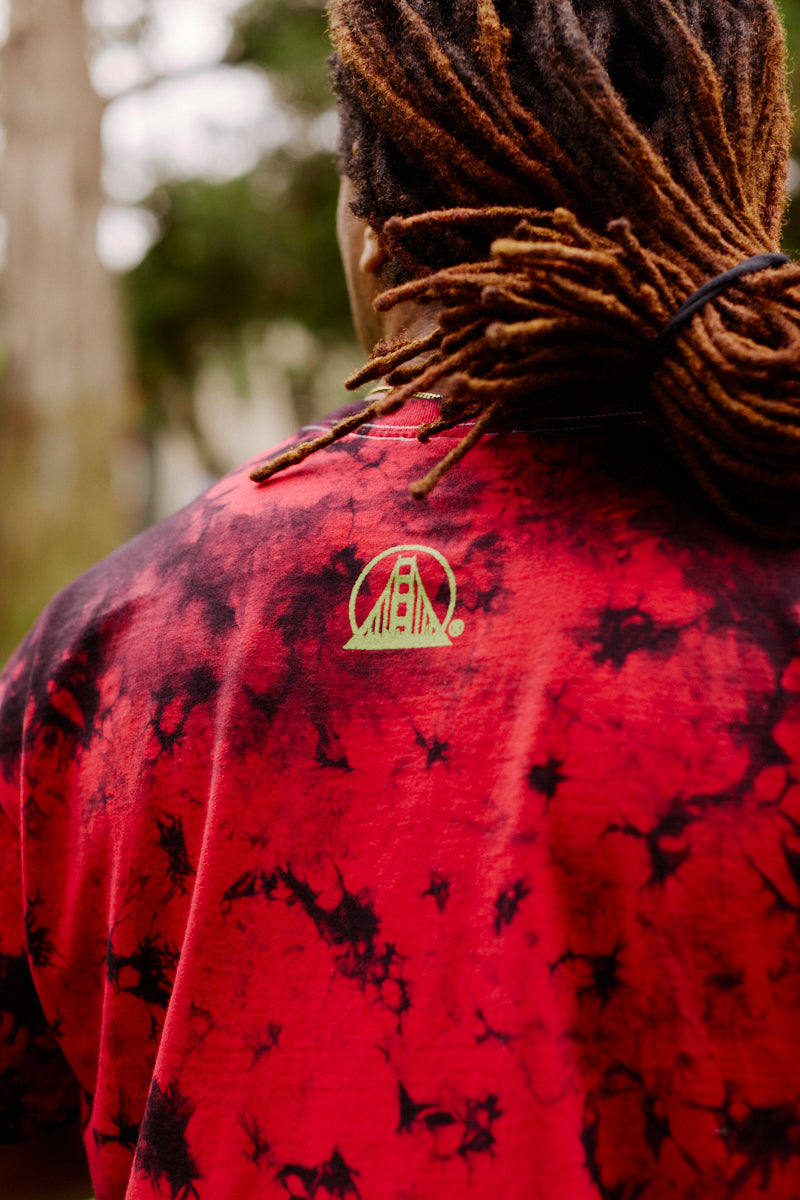 A person with dreadlocks wears the Gold Rush Tee - Red Crystal Dye, a unisex red tie-dye shirt featuring a yellow bridge logo on the back, outdoors—perfect for any 49ers or Gold Rush fan.