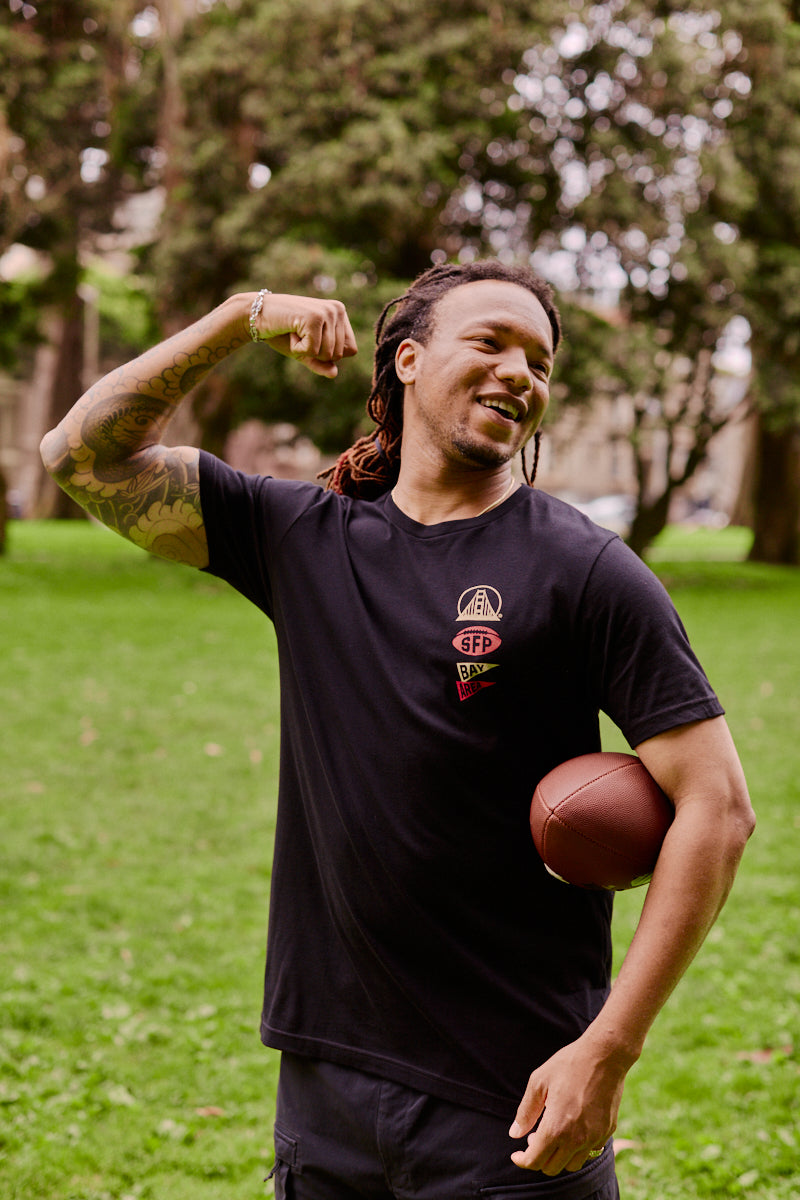With a football tucked under one arm and his other arm flexed in the park, a smiling man with dreadlocks proudly sports the Niners Faithful Tee—perfect for showing off that San Francisco spirit.