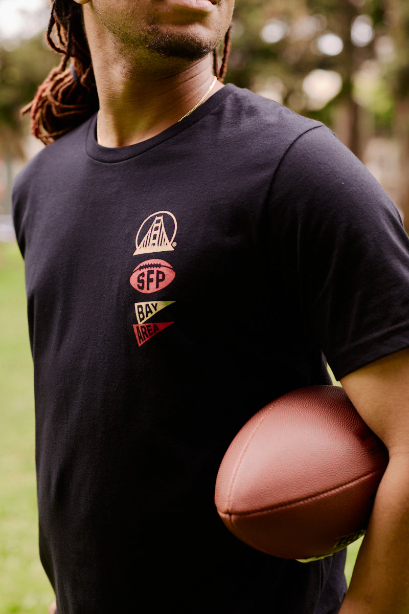 A person stands outdoors on the grass, holding a football under their arm and wearing the Niners Faithful Tee—a unisex black shirt perfect for showing off your San Francisco pride.