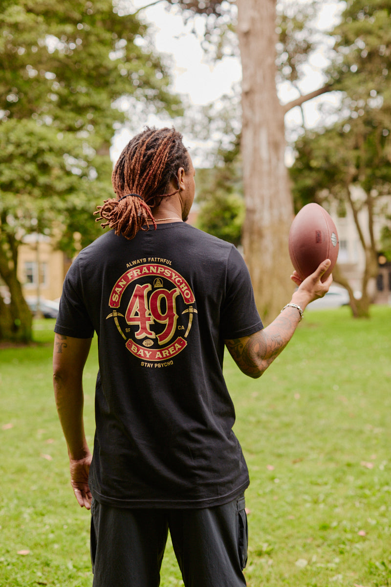 Wearing the Niners Faithful Tee, a person with dreadlocks holds a football while standing in a grassy park in San Francisco.