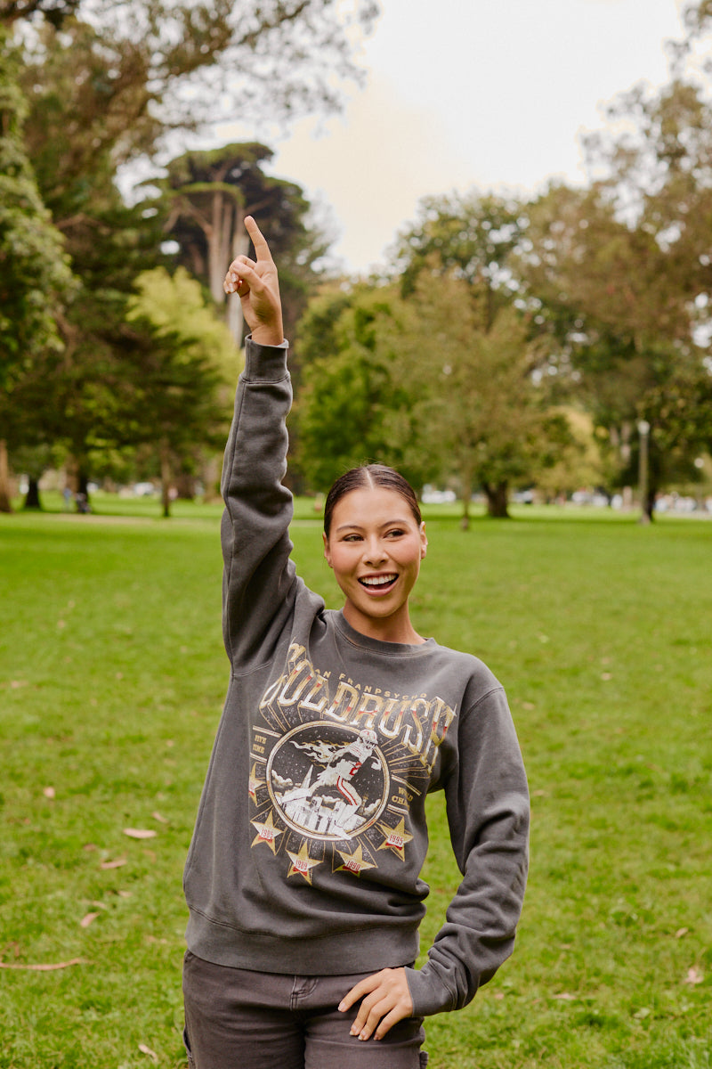 A smiling person wearing the Gold Rush Crewneck stands on grass in a park, raising one arm and pointing upward.