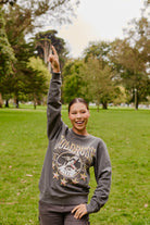 A smiling person wearing the Gold Rush Crewneck stands on grass in a park, raising one arm and pointing upward.