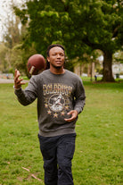 A man wearing the Gold Rush Crewneck tosses a football while walking on green grass in a park.