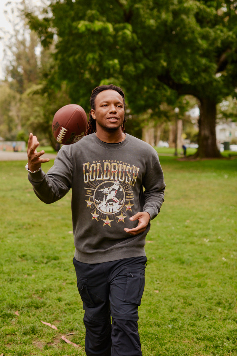 A man wearing the Gold Rush Crewneck tosses a football while walking on green grass in a park.