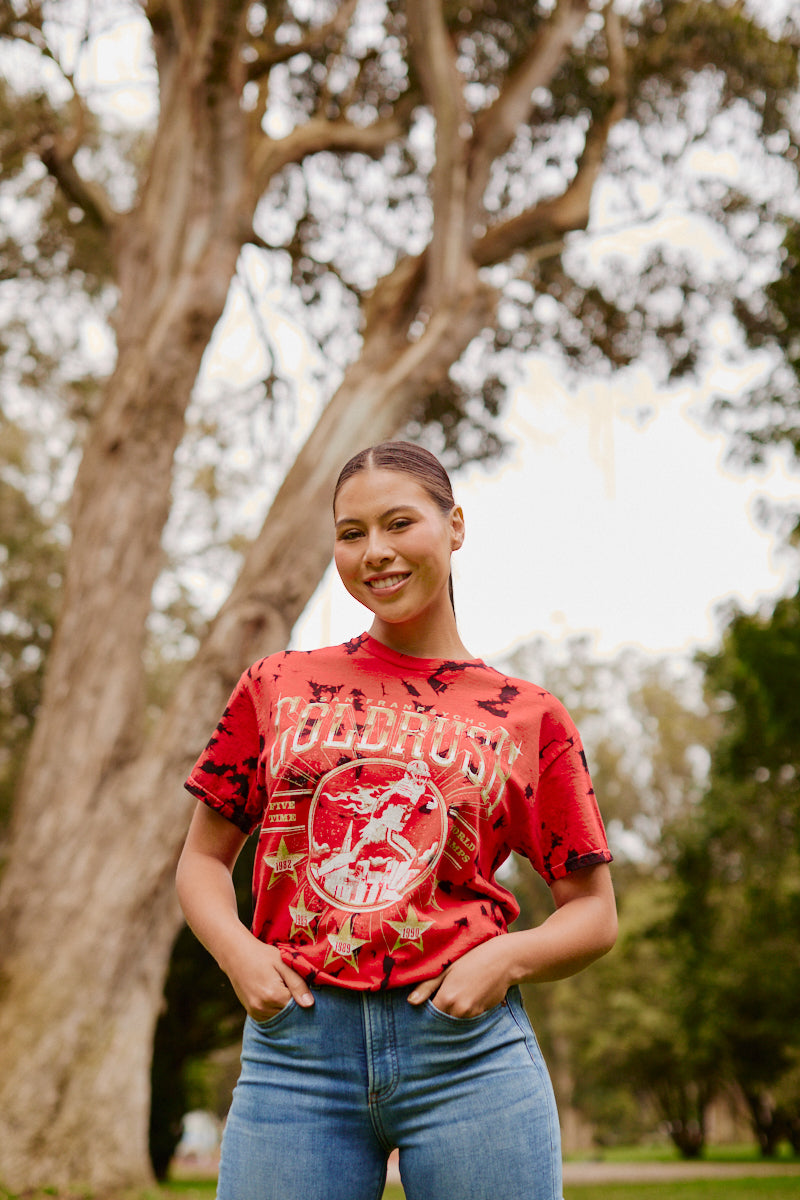 A woman smiling outdoors in the Gold Rush Tee - Red Crystal Dye and jeans, standing in front of trees in a park.
