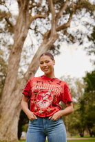 A woman smiling outdoors in the Gold Rush Tee - Red Crystal Dye and jeans, standing in front of trees in a park.