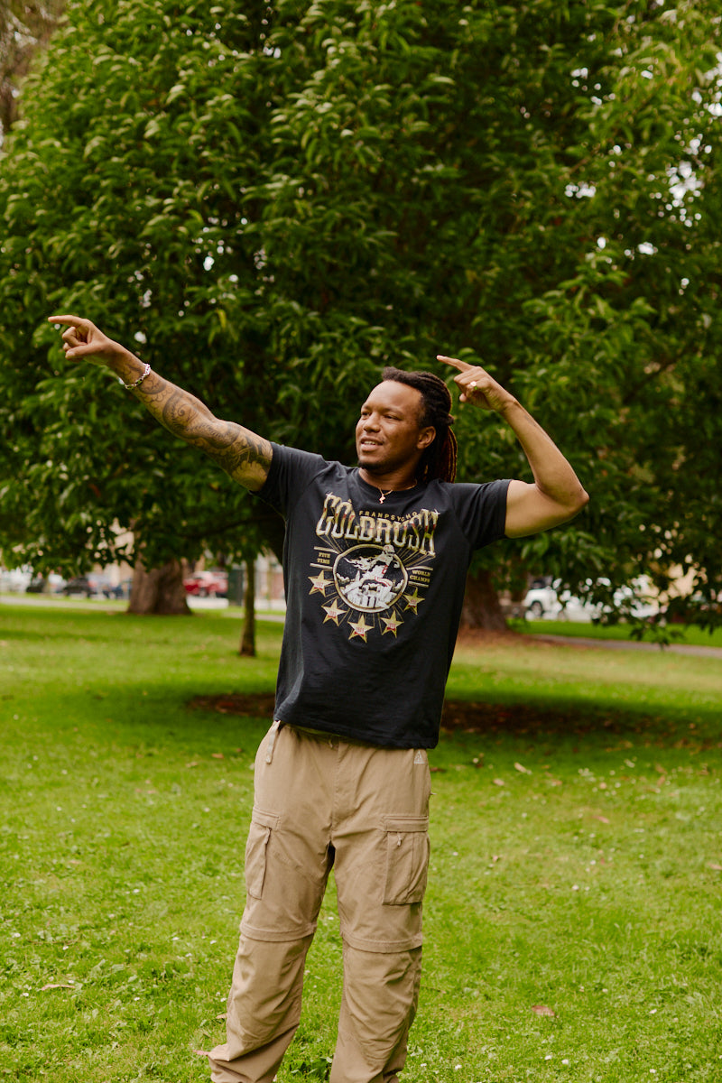 A person stands on grass, pointing to the side and looking up, wearing the Gold Rush Tee in Vintage Black with trees and greenery in the background—a classic San Francisco style.