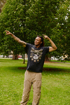 A person stands on grass, pointing to the side and looking up, wearing the Gold Rush Tee in Vintage Black with trees and greenery in the background—a classic San Francisco style.
