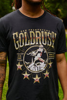A man wearing the Gold Rush Tee - Vintage Black, featuring a football graphic and championship years, celebrates the spirit of San Francisco.