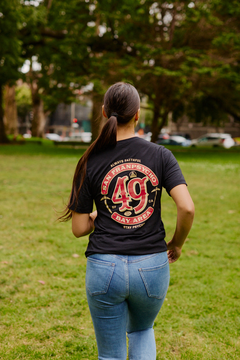 A woman with a ponytail walks on grass, wearing the Niners Faithful Tee with a classic unisex fit; trees are visible in the background.
