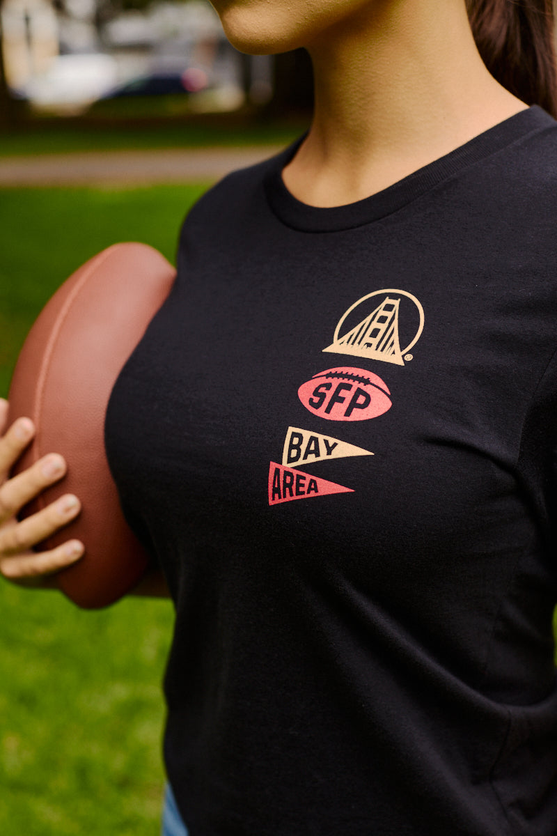 Person holding a football, wearing the Niners Faithful Tee—a black unisex fit shirt featuring SFP Bay Area and a bridge graphic on the front—making it an ideal addition to any San Francisco Niners fan collection.