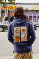 A person with dreadlocks wears the Sunset Dahlia Pullover Hoodie, a vintage mineral wash hoodie adorned with Japanese art-inspired graphics of the Golden Gate Bridge, waves, sun, and a flower—echoing the design found on the Sunset Dahlia Tee.