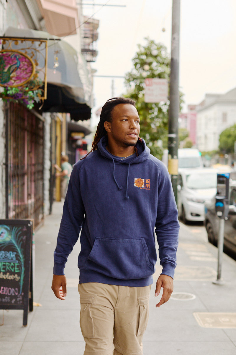 A man in a blue Sunset Dahlia Pullover Hoodie and tan pants walks on a city sidewalk near shops and parked cars, sporting a Japanese art-inspired tee with a vintage mineral wash, hand printed in San Francisco.
