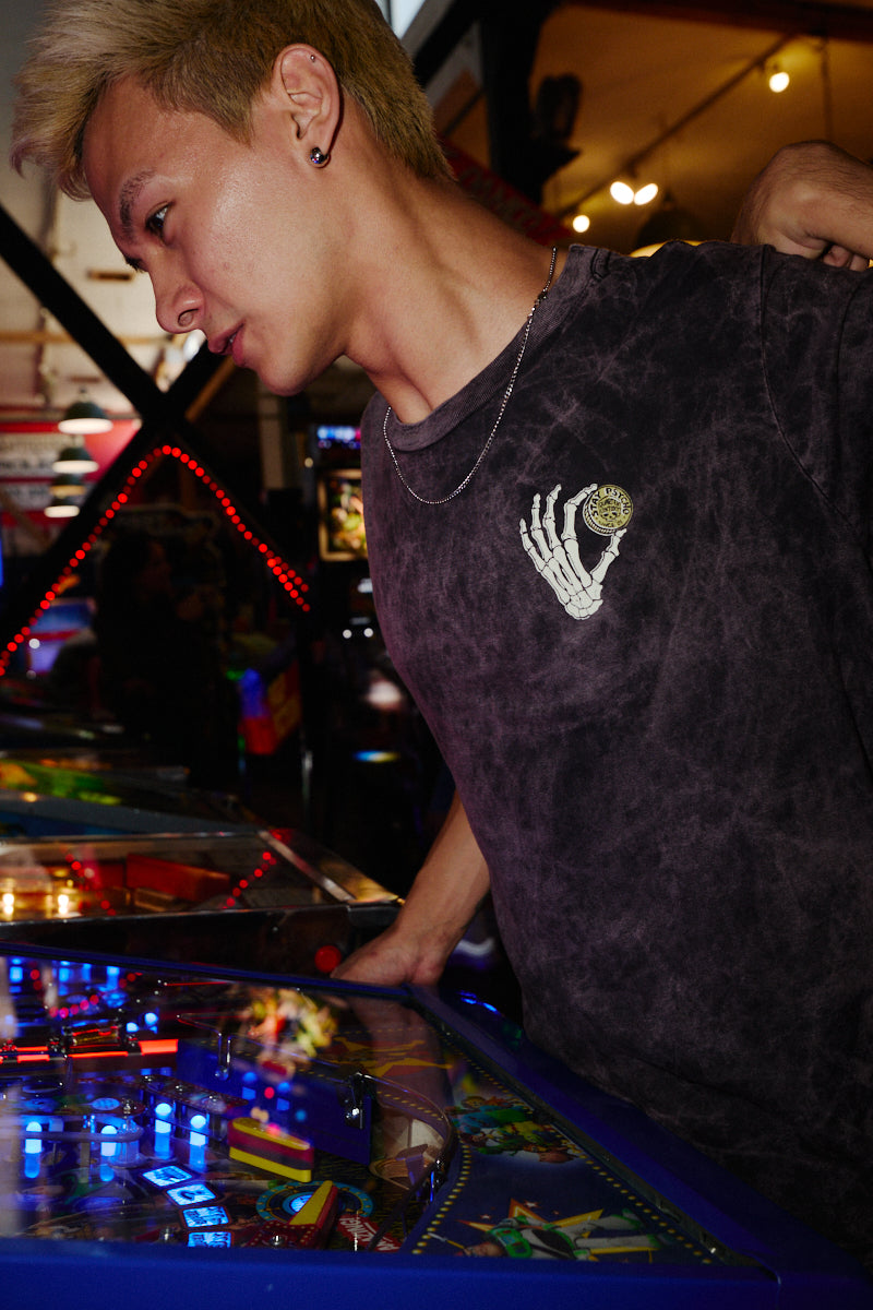 Person wearing a Quarter Cent Tee with an acid-wash skeleton hand graphic while playing a pinball machine.