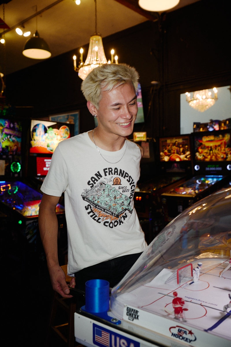 A person smiles at a tabletop hockey game wearing a white Still Cookin’ Tee with a San Francisco illustration.