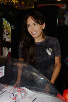 A smiling woman in a grey Quarter Cent Tee with a skeleton hand graphic plays an arcade game.