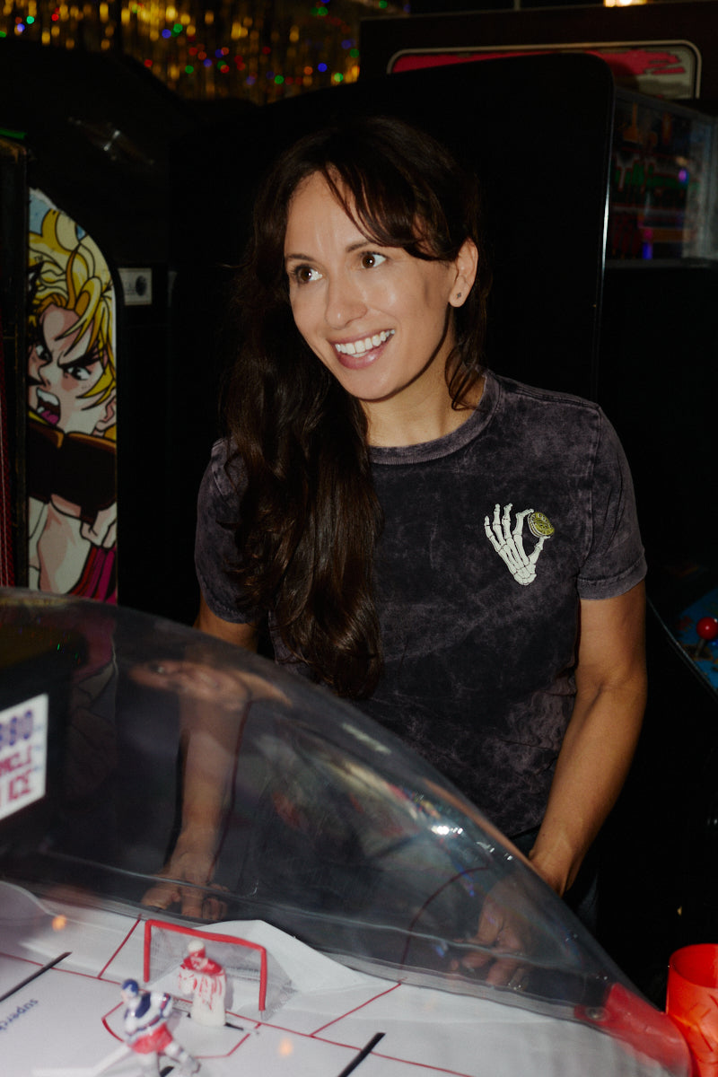 A smiling woman in a grey Quarter Cent Tee with a skeleton hand graphic plays an arcade game.