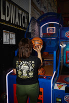 A woman in a black Women’s Quarter Cent Tee holds a basketball in front of an arcade game.