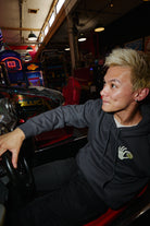 Person wearing a dark grey Quarter Cent Pullover Hoodie with a skeleton hand graphic, sitting in a racing arcade game.