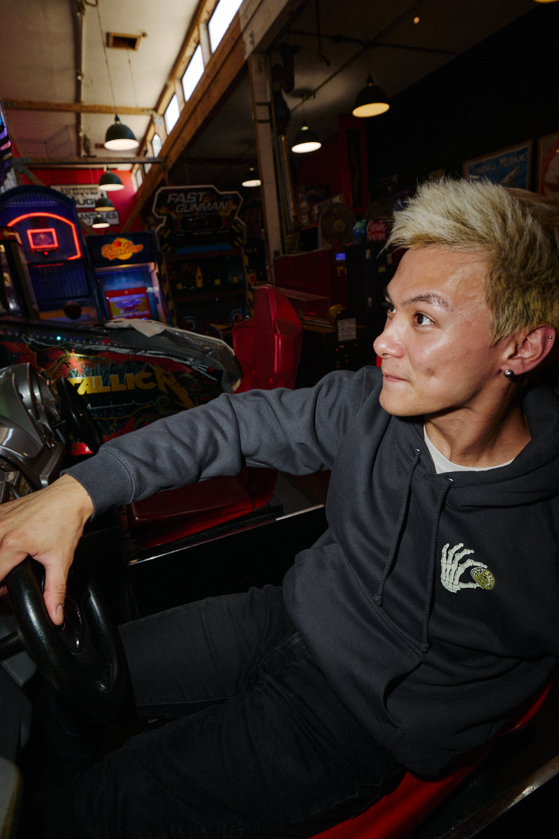 Person wearing a dark grey Quarter Cent Pullover Hoodie with a skeleton hand graphic, sitting in a racing arcade game.