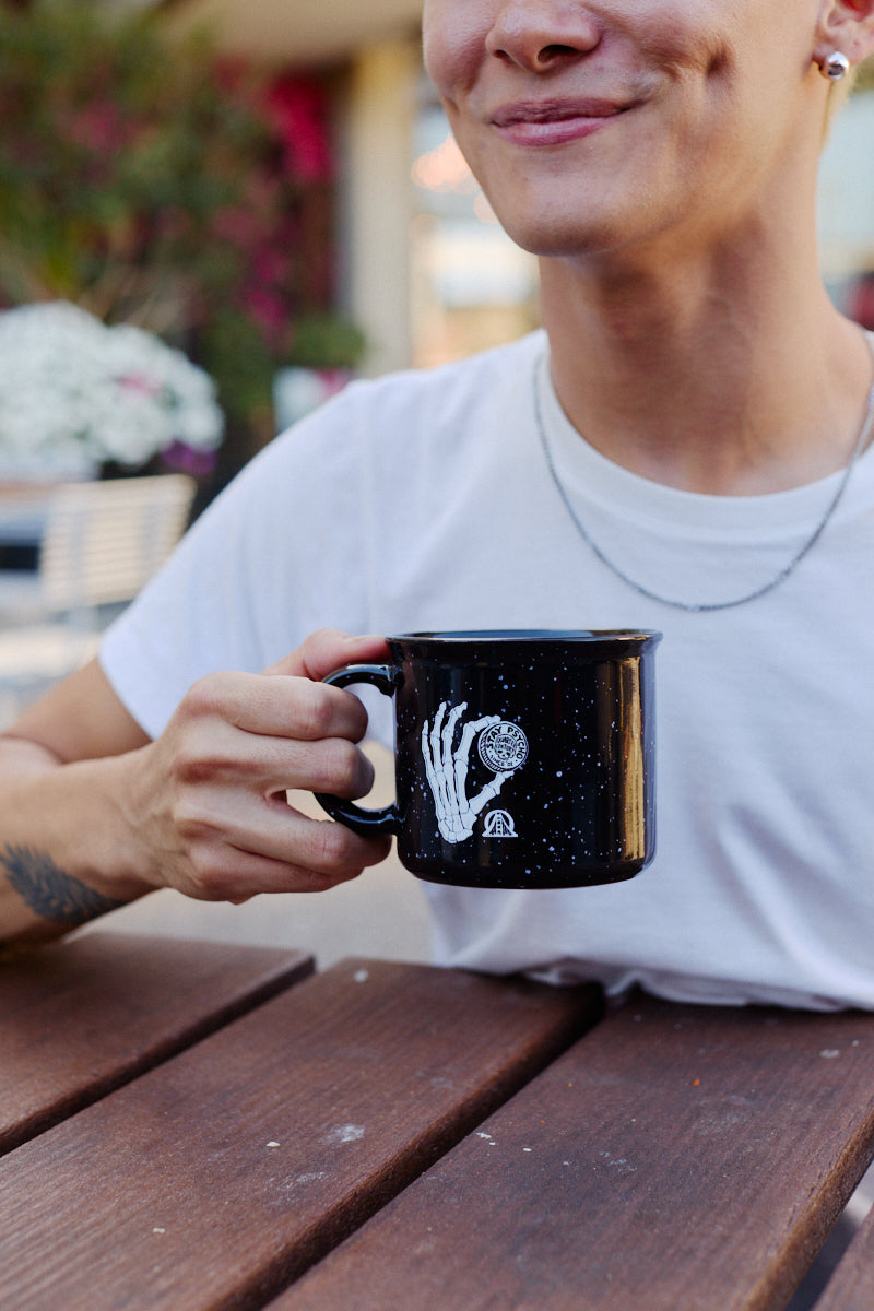 A person holds a speckled Quarter Cent Mug featuring a white skeletal hand graphic.