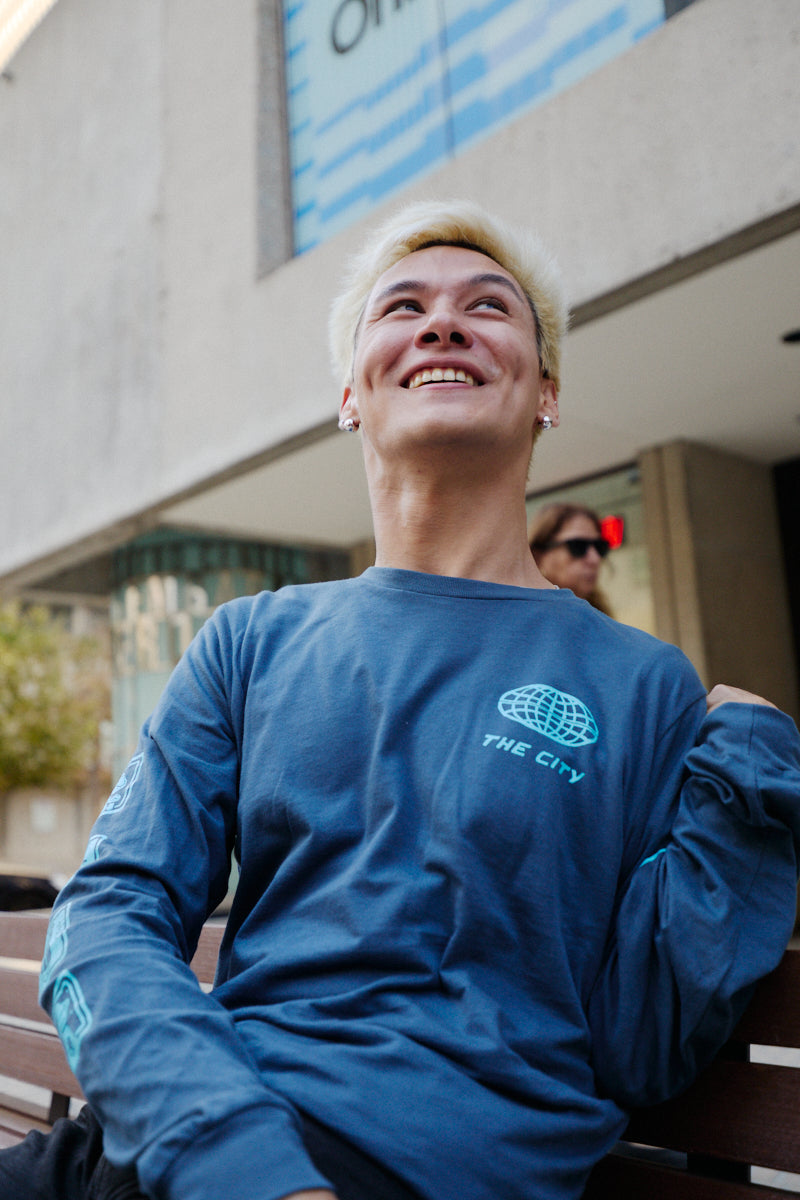 A person wears a blue 3D Logo Long Sleeve featuring a globe graphic and THE CITY text on the chest.