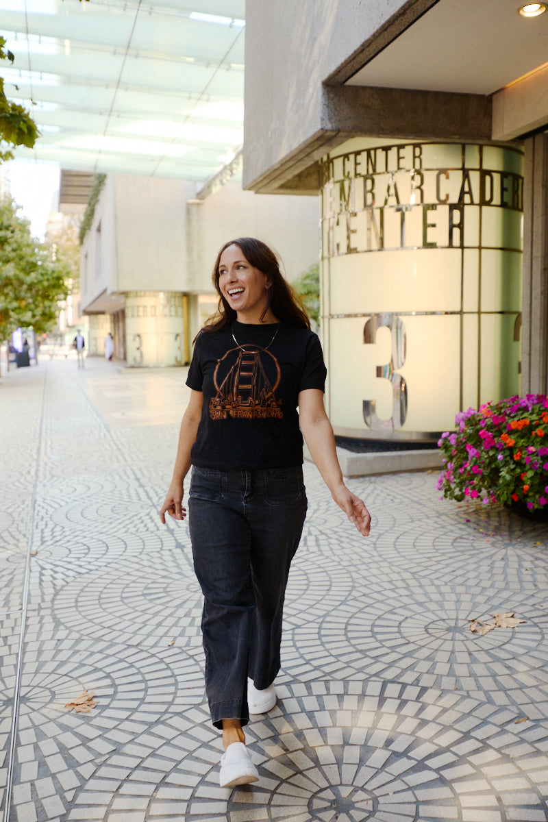 A smiling woman wears a Women’s 3D Logo Tee with a retro-futurism Golden Gate Bridge graphic while walking outdoors.