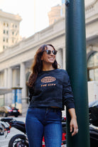 Woman wearing a Women’s The City Black Camo Crewneck crop hoodie with an orange graphic that reads THE CITY.