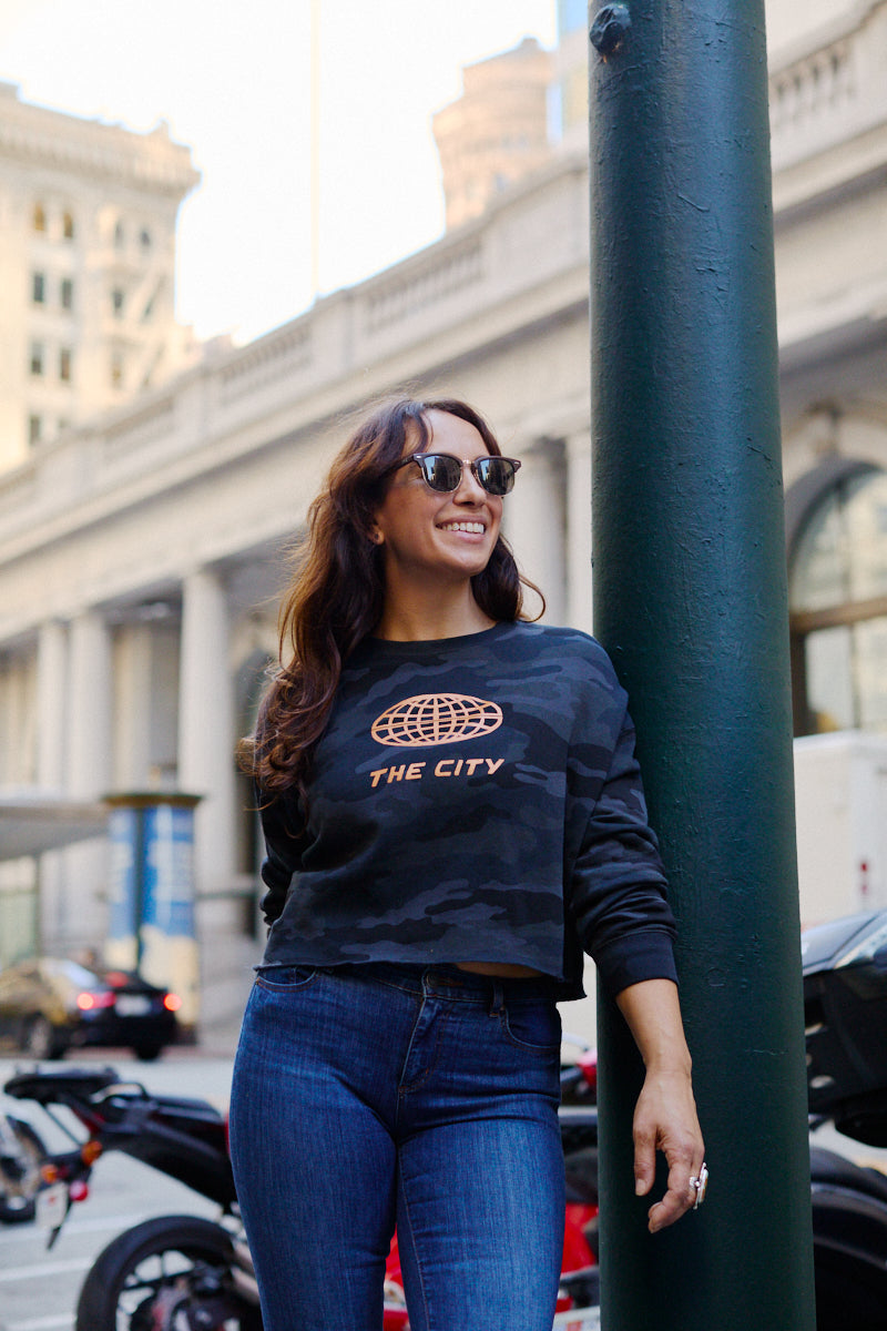 Woman wearing a Women’s The City Black Camo Crewneck crop hoodie with an orange graphic that reads THE CITY.