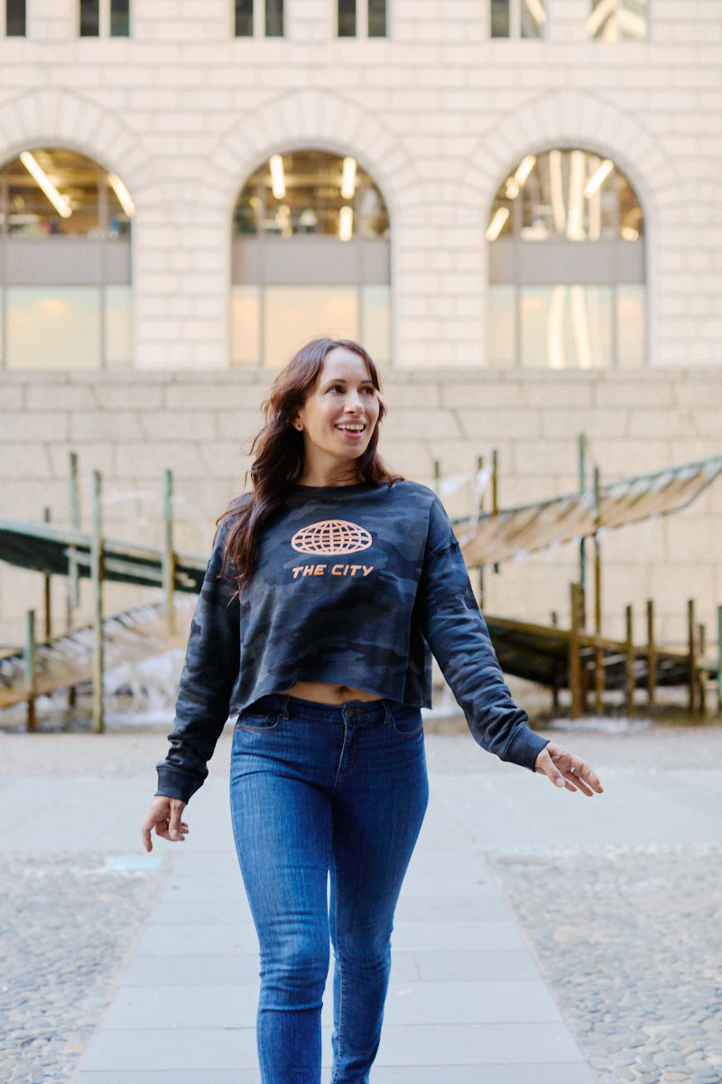 A woman wears a Women’s The City Black Camo Crewneck, a black camo crop sweatshirt with a globe graphic and THE CITY.