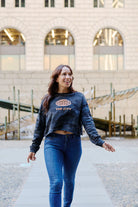 A woman wears a Women’s The City Black Camo Crewneck, a black camo crop sweatshirt with a globe graphic and THE CITY.