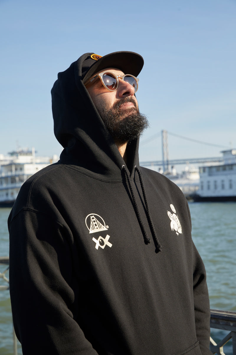 A bearded man in a black Moon Bridge Pullover Hoodie and sunglasses gazes upward by the water, with boats and a bridge behind him.