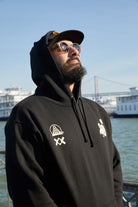 A bearded man in a black Moon Bridge Pullover Hoodie and sunglasses gazes upward by the water, with boats and a bridge behind him.