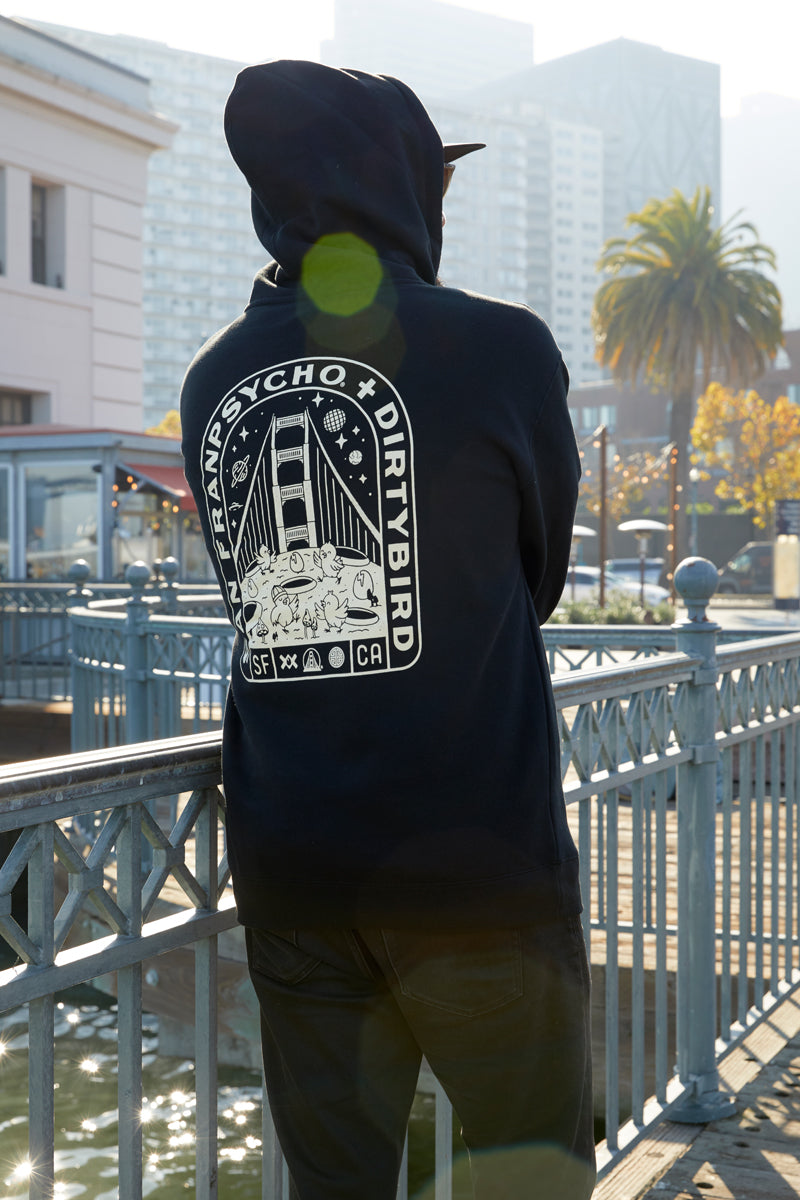 Wearing the Moon Bridge Pullover Hoodie, a person stands by a railing with city buildings and a palm tree in the background.