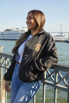 A woman in a Dirty UFO Coaches Jacket stands by a waterfront railing with boats and a bridge behind her, highlighting Bay Area streetwear style.