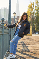 A woman wearing the Dirty UFO Coaches Jacket and ripped jeans leans on a railing outdoors with city buildings behind her, capturing a true Bay Area streetwear vibe.