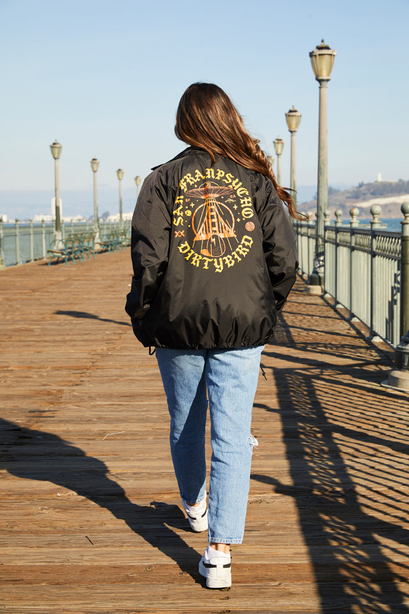 A woman walks on a wooden boardwalk, wearing the Dirty UFO Coaches Jacket from the SFP x Dirtybird collab with blue jeans—a stylish mix of Bay Area streetwear flair.