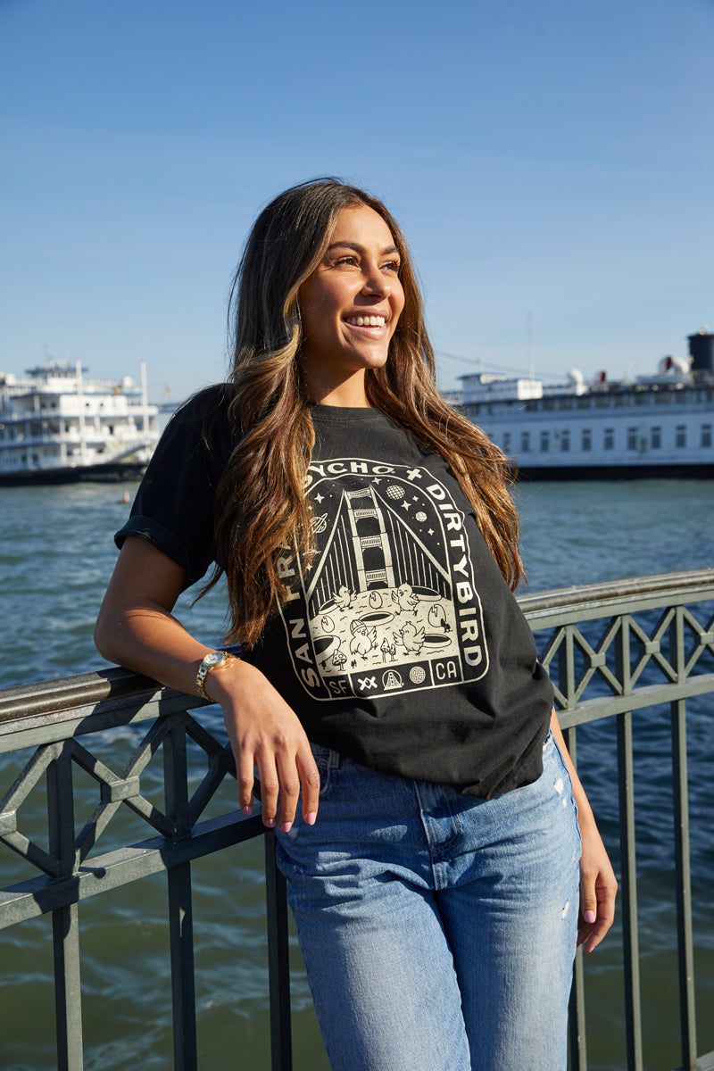 A woman in a Moon Bridge Tee leans on a waterfront railing in San Francisco, with boats in the background.
