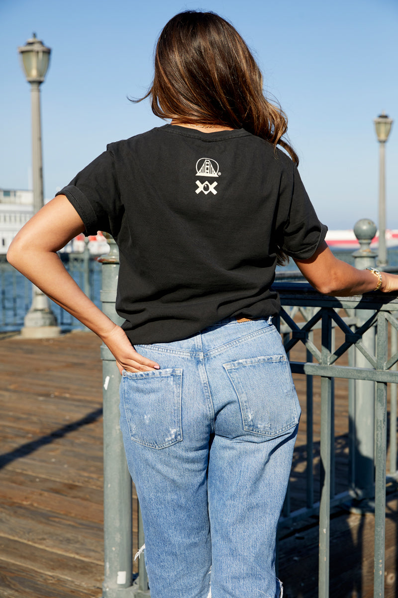 A woman with long hair stands on a San Francisco pier, back to the camera, wearing the Moon Bridge Tee in black and blue jeans.