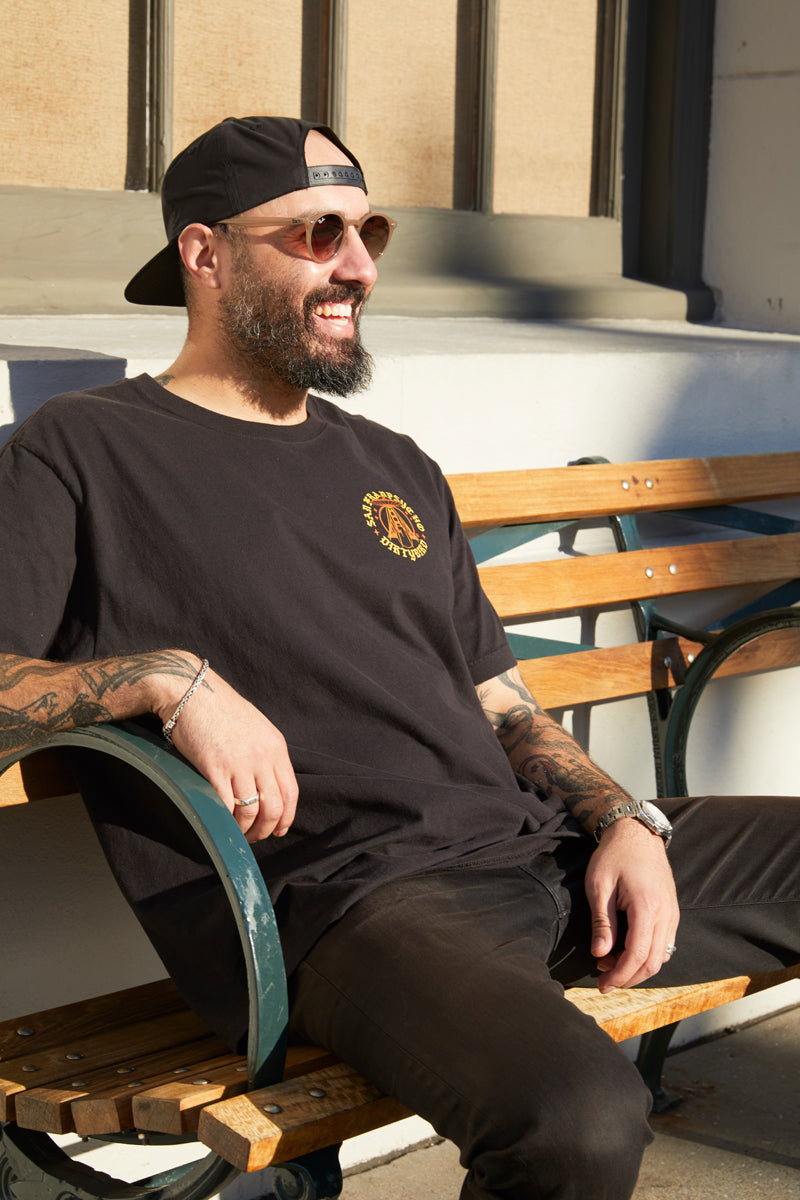 Man with a beard and sunglasses smiles while sitting on a bench in the sunlight, sporting the Dirty UFO Tee and a cap—a perfect nod to Bay Area culture.