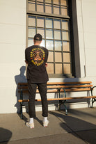 A person in a black Dirty UFO Tee and cap stands by a bench outside, facing a sunny window—a scene reflecting Bay Area culture and the creative spirit behind San Francisco hand-printed apparel.