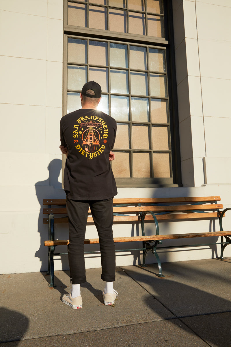 A person in a black Dirty UFO Tee and cap stands by a bench outside, facing a sunny window—a scene reflecting Bay Area culture and the creative spirit behind San Francisco hand-printed apparel.
